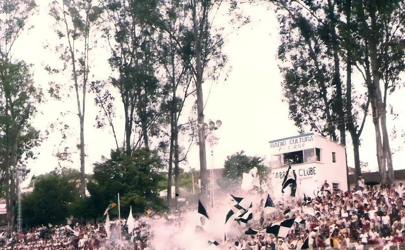 Torcida do Fabril com bandeiras pretas e brancas e fumaça na arquibancada, diante da cabine da Rádio Cultura