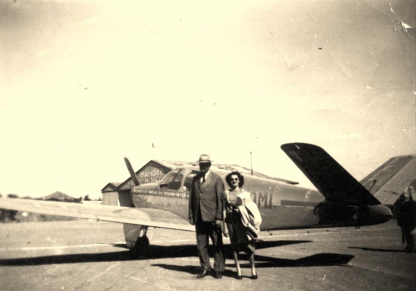 Um casal posa diante de um avião no antigo aeroporto de Lavras, com o Hangar Vaz Monteiro ao fundo